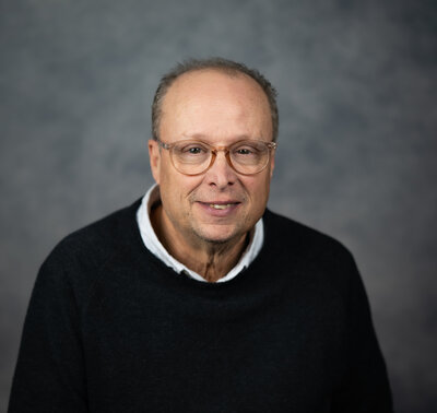 John Koon, a man with gray hair and glasses, smiling in a dark sweater over a white collared shirt.