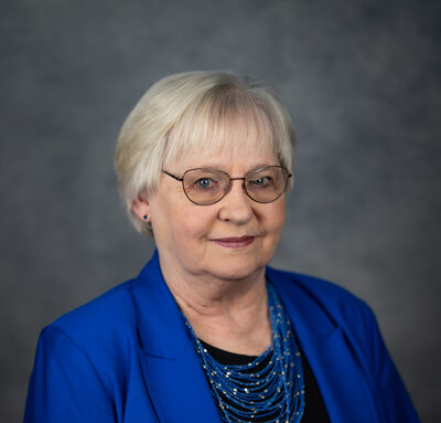 Karon Beukema, a woman with short gray hair and glasses, smiling in a bright blue jacket.