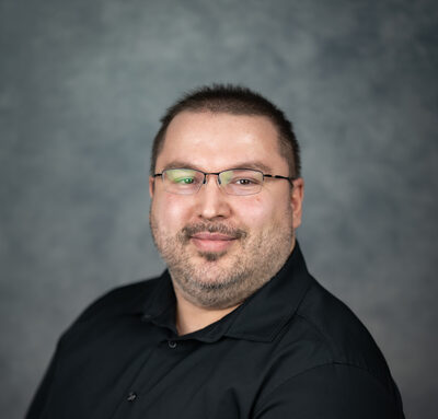 Shawn Arapovich, a man with short dark hair and glasses, smiling in a black shirt.